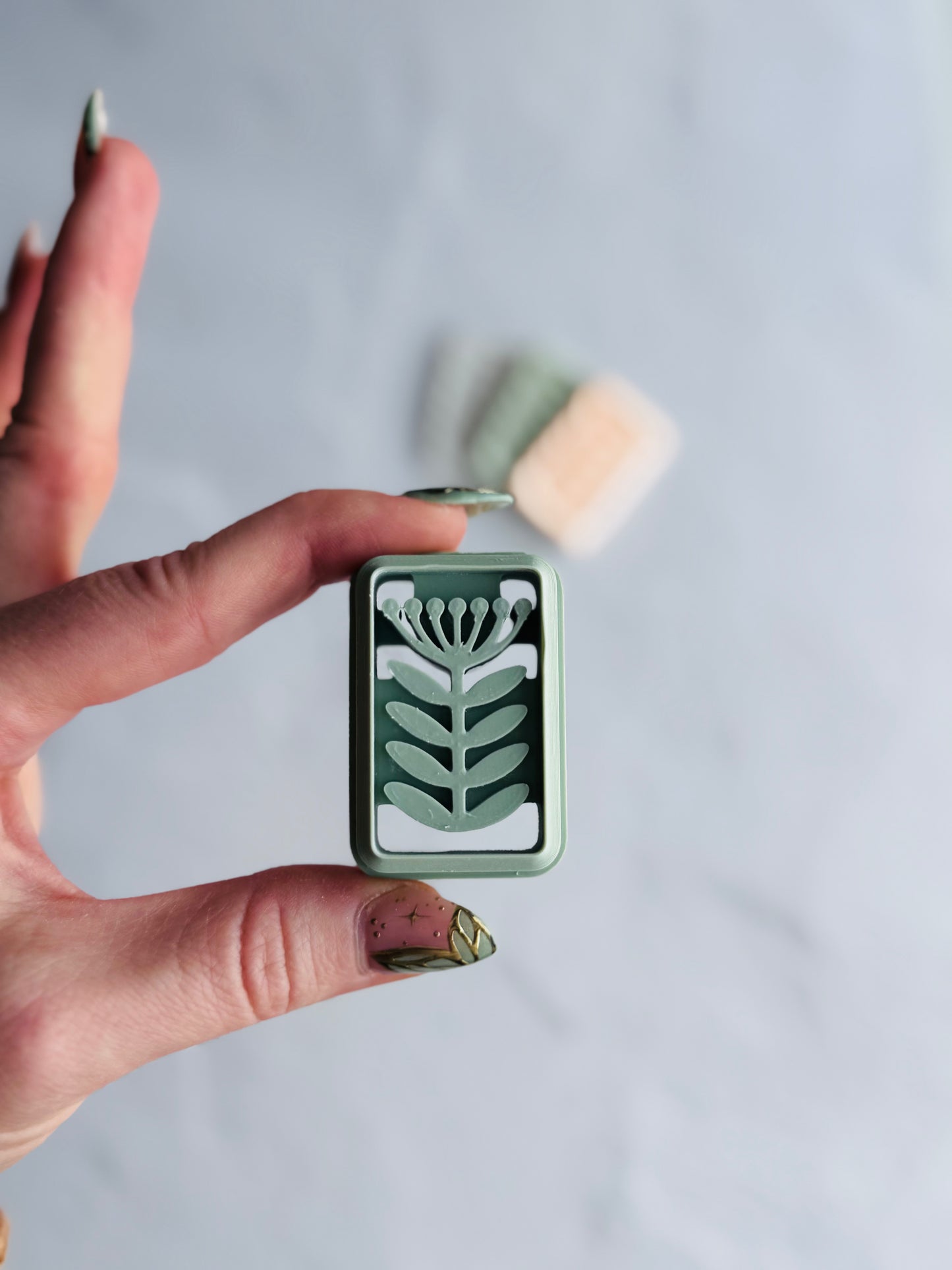 Hand holding a small green polymer clay cutter with floral design against a light gray background